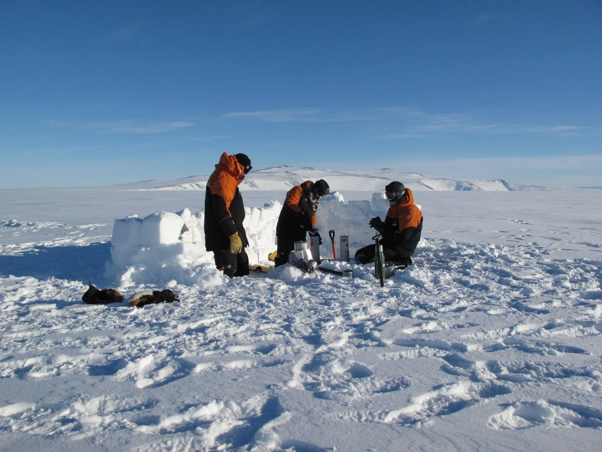 Three team members working behind a low wall of snow blocks on the Antarctic ice.