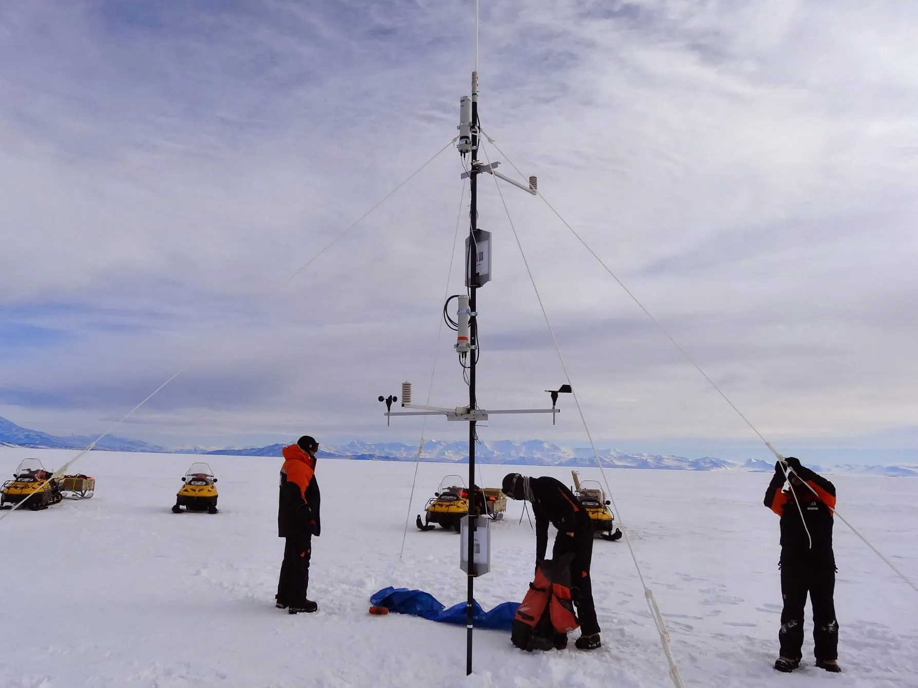 An automatic weather station on a guyed mast with wind and temperature sensors, the field team and snowmobiles around it on the Antarctic ice.