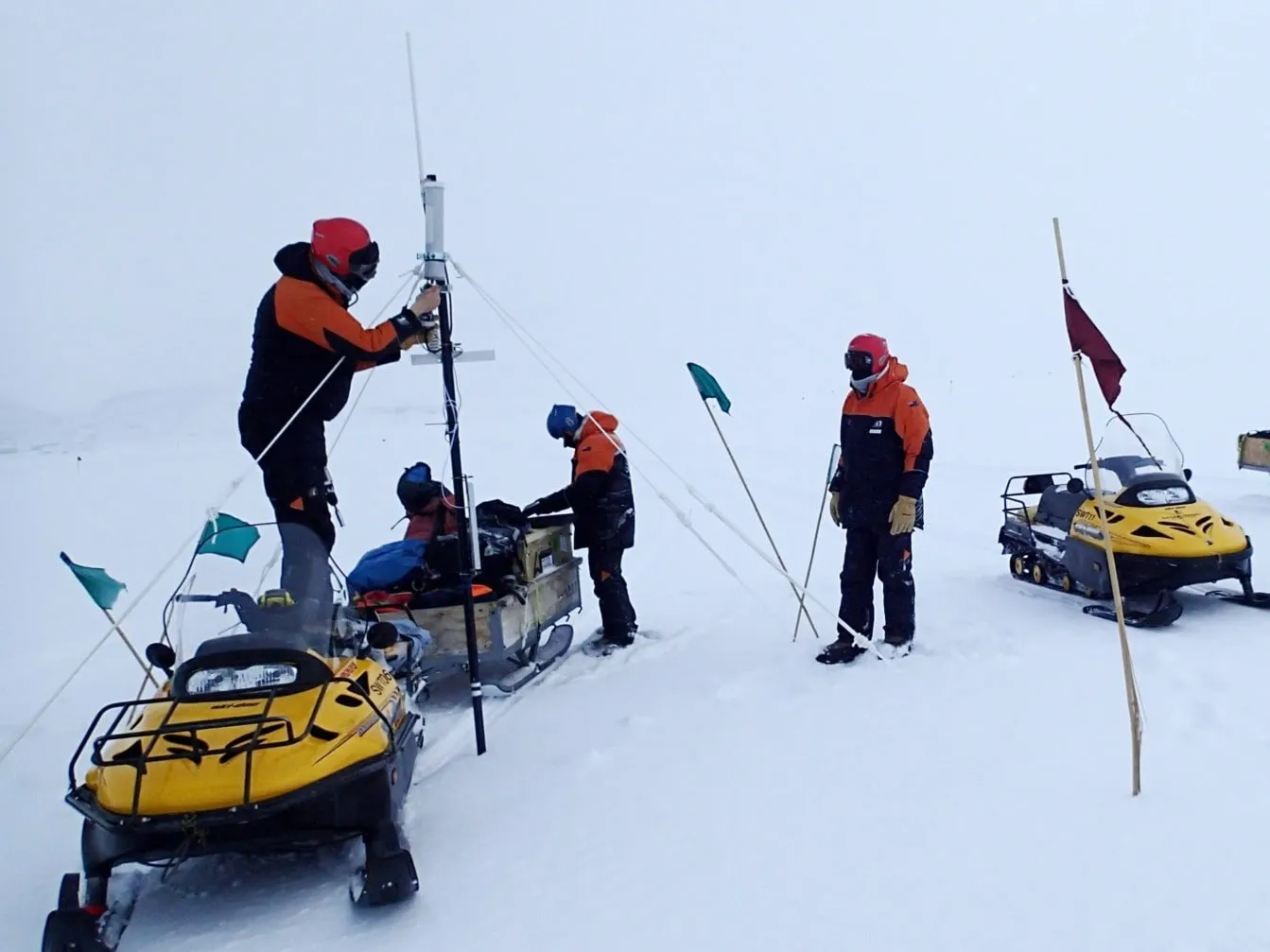 A field team in polar gear installing a weather station mast beside snowmobiles in poor visibility.